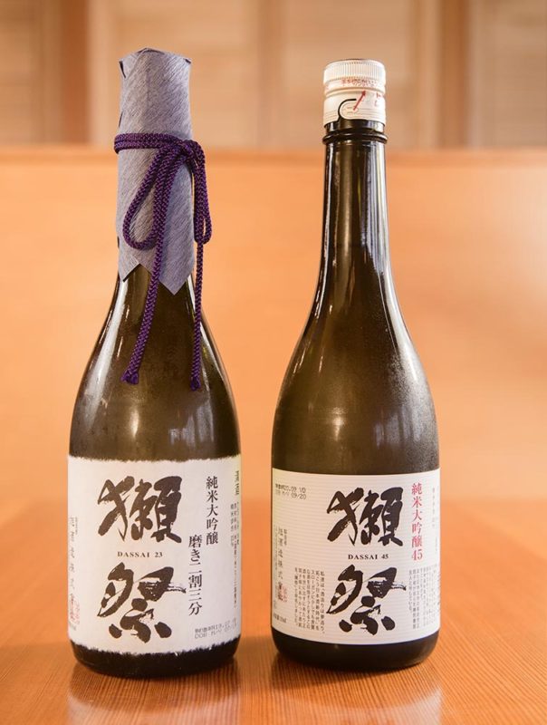 Two bottles of Dassai sake in a wooden room.
